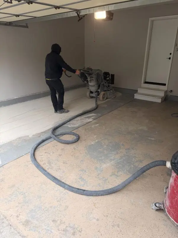 Crew member diamond grinding an old coating off a garage floor, with a dust vacuum hose across the slab