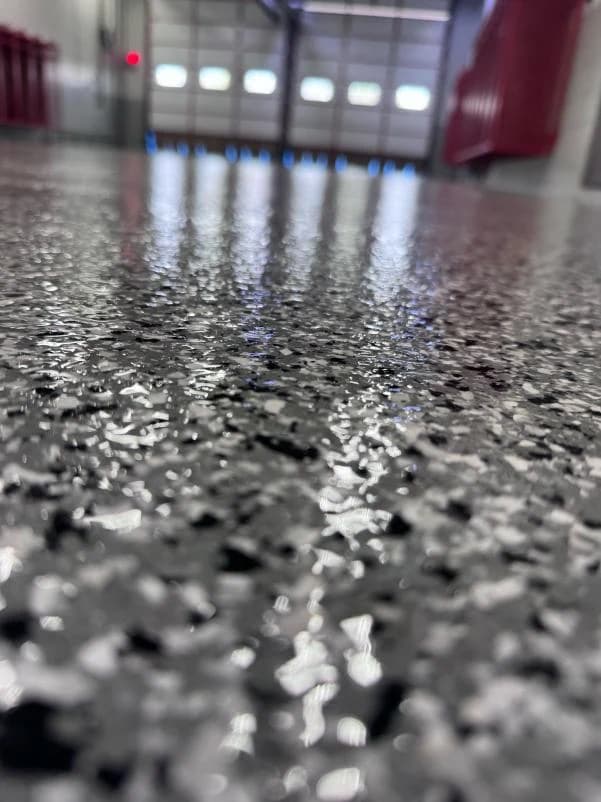 Close-up of a gray, black and white flake floor coating under a clear topcoat, with overhead doors out of focus behind it