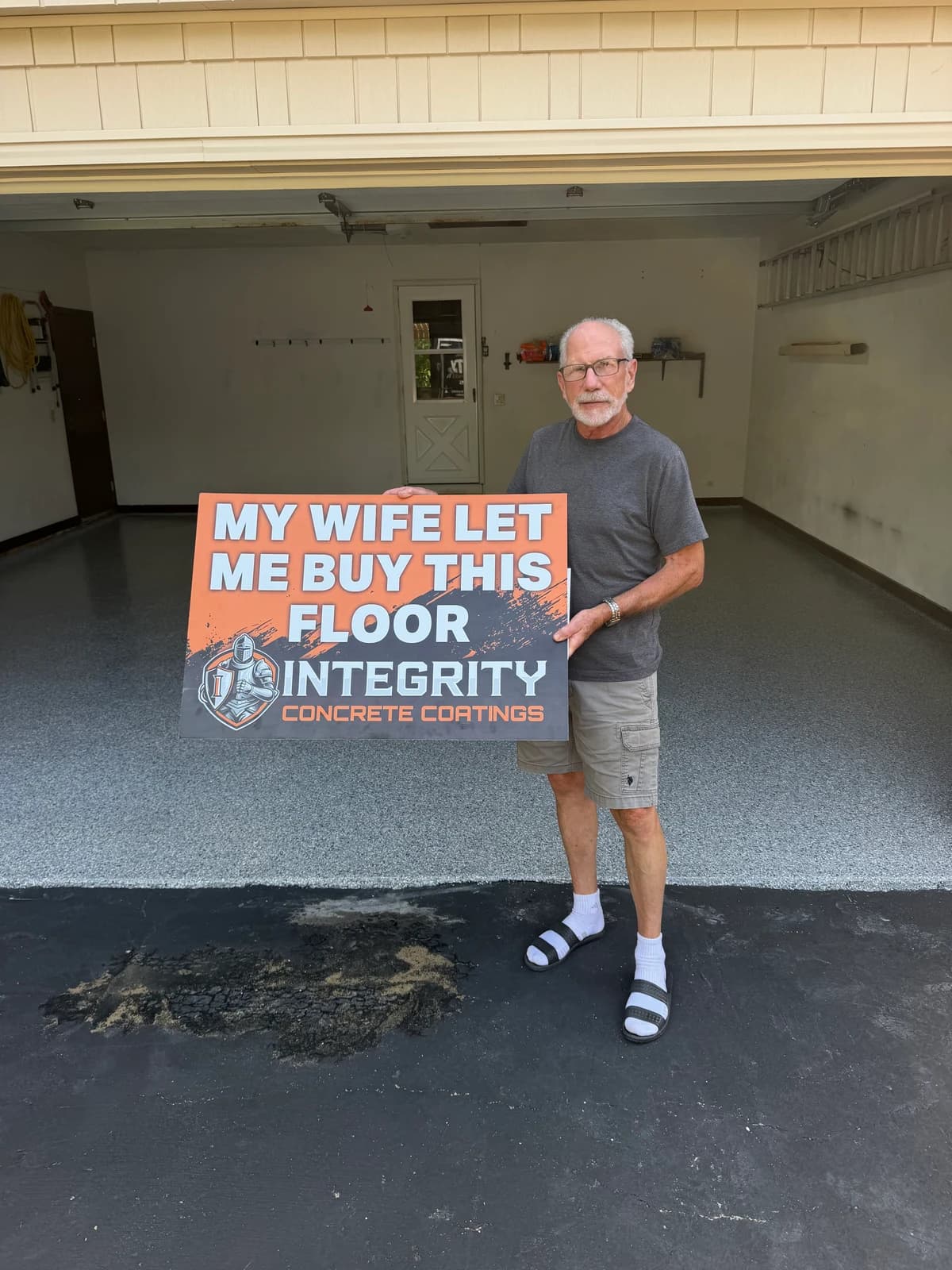 Homeowner holding a My Wife Let Me Buy This Floor sign in a freshly coated garage