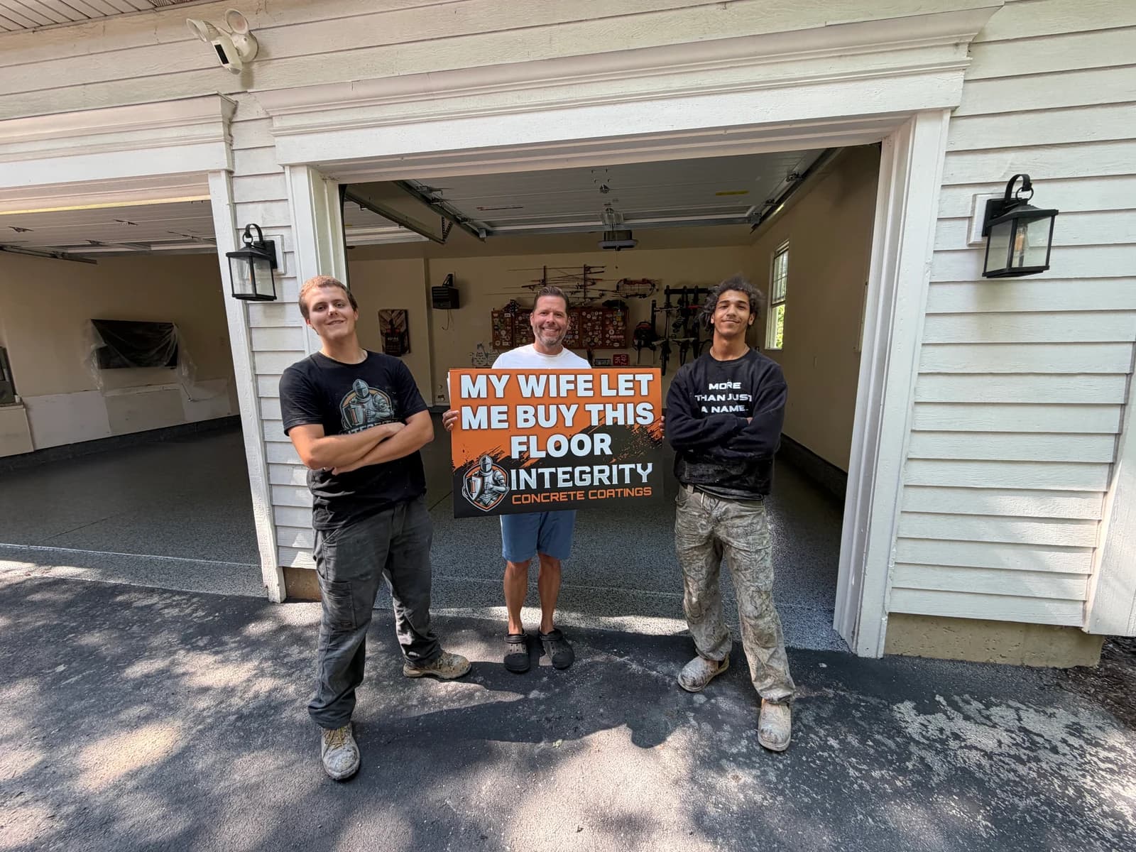 Integrity crew and homeowner with a My Wife Let Me Buy This Floor sign at a two car garage