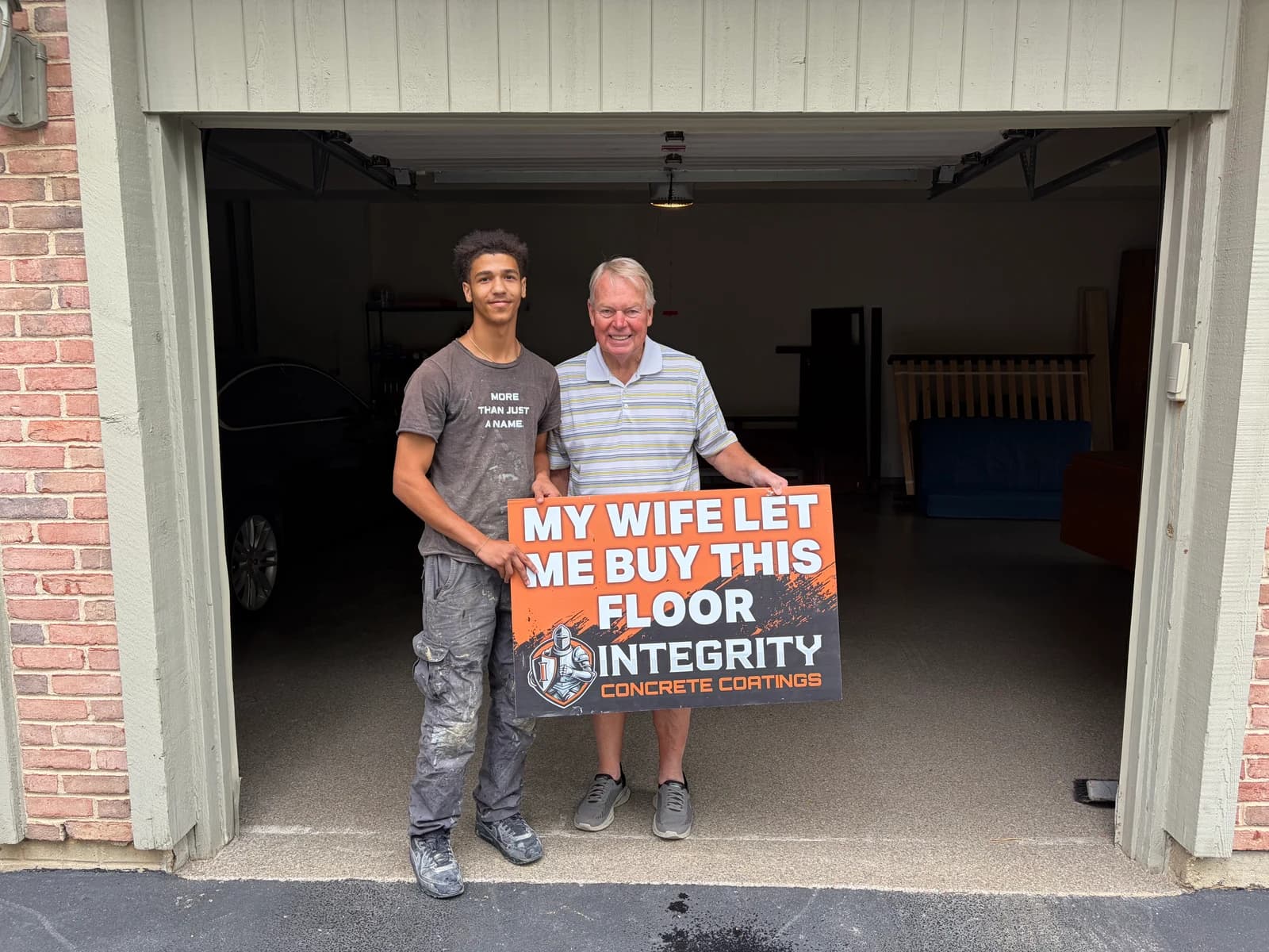 Crew member and homeowner with a My Wife Let Me Buy This Floor sign at a brick home garage
