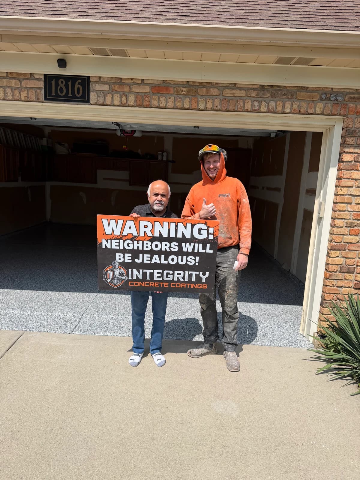 Homeowner and crew member holding a Neighbors Will Be Jealous sign outside a brick home garage