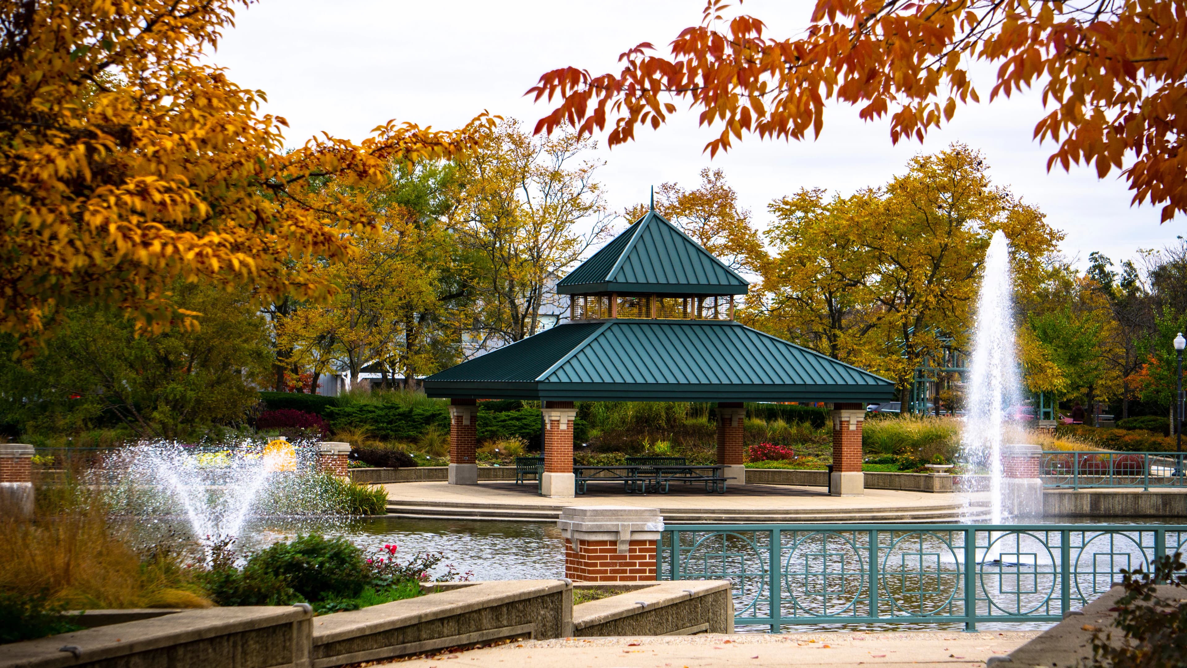Green-roofed pavilion on an island in a park pond, with fountains and fall foliage around it