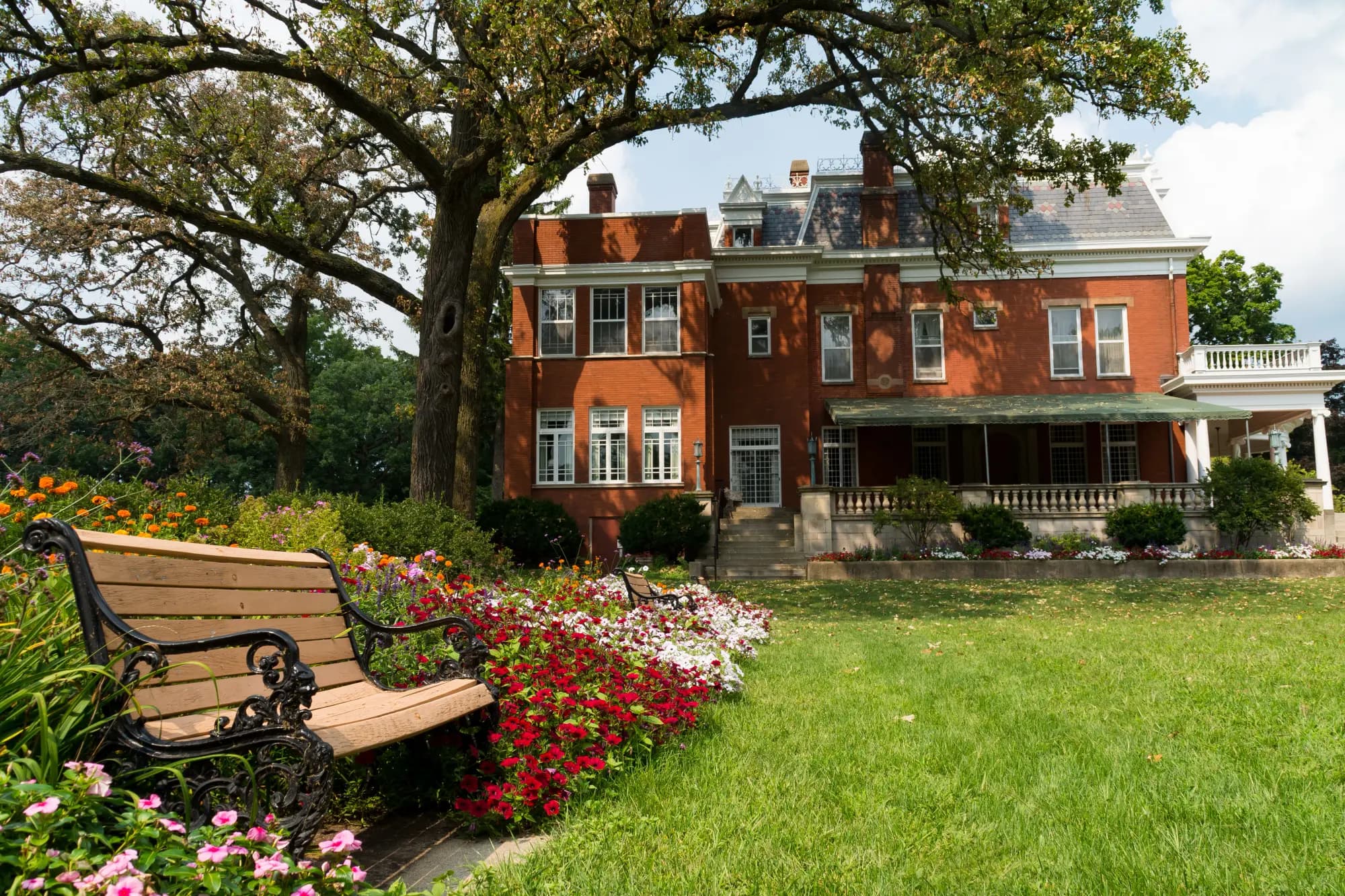 Three-story red brick Victorian mansion behind flower beds and a lawn, with a park bench under a large oak