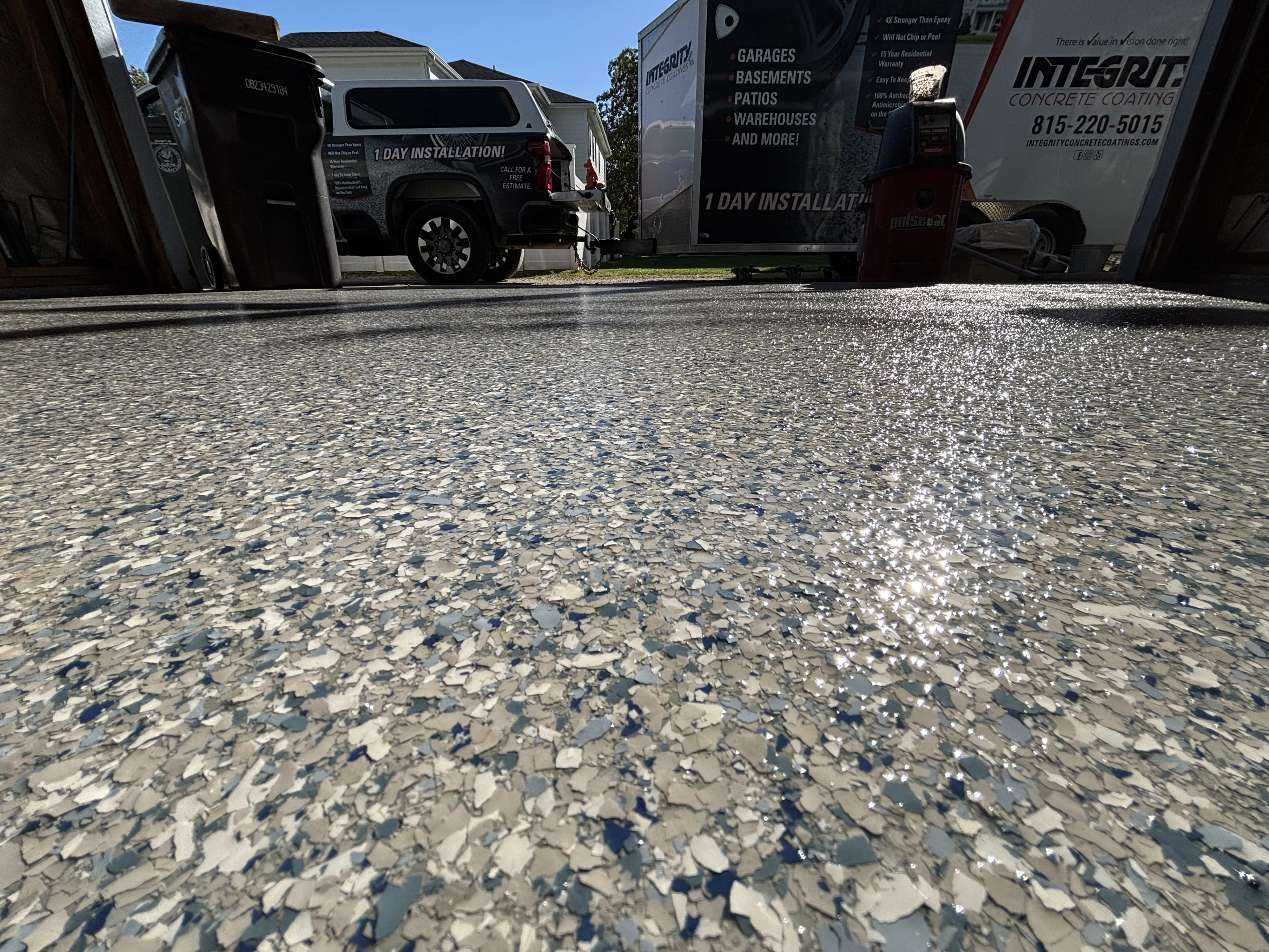 Low-angle view of a glossy blue, gray and white flake garage floor coating, with an Integrity Concrete Coatings truck and trailer parked outside